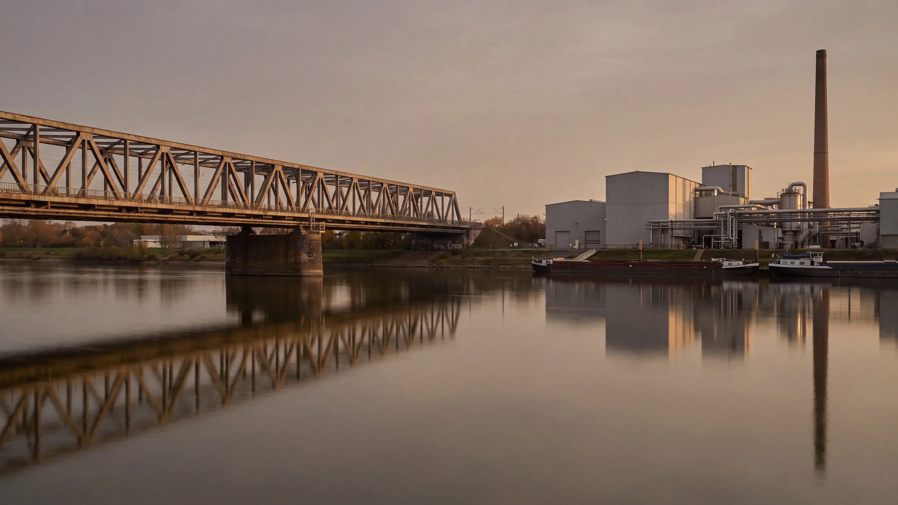 Eine Straßenbrücke über einen Fluss, dahinter die Anlagen eines Industriewerks, gespiegelt im Wasser.